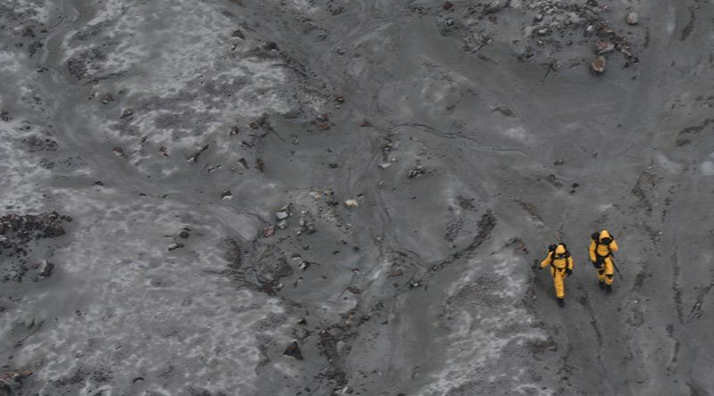 Two New Zealand Defence Force explosives-ordnance disposal experts in hazmat suits set out to recover bodies from the White Island volcano debris field. NZDF photo.