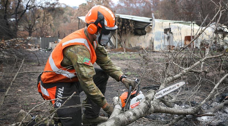 Private Jake Jubelin, an Army Reserve infantryman from the 2nd/17th Battalion, Royal NSW Regiment, cuts apart a fallen tree to restore access to a burn-out property. The owner’s home (background) was destroyed by bushfire, with the elderly couple forced to share a tent until access for a new caravan was restored by the Army. Photo by Major Cameron Jamieson