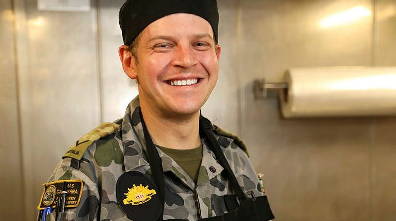 Lance Corporal Christopher Lyne puts on his apron on before preparing meals in HMAS Canberra's galley for the Ship's Company and embarked forces on the way to Exercise Talisman Sabre 21. Photo by Leading Seaman Nadev Harel.